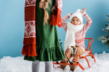 selective focus of boy in winter hat sitting with hands above head on sleigh near sister on blue