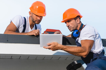 Wall Mural - Selective focus of handymen in workwear using digital tablet while working on roof of building