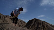 Barefoot Male Musician Plays A Wind Instrument Saxophone In The Mountains Against The Sky