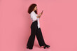 Full length profile photo of charming redhead lady hold phone in hands, walking street, browsing smartphone addicted user wear casual clothes isolated on pink background