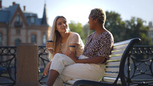 Two Multiethnic Female Sitting On Bench Outdoors Drinking Coffee Together.