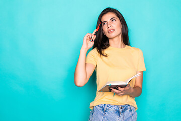 Pretty young beautiful woman standing, writing, take notes, holding textbook notebook organizer in hand and pen on turquoise background