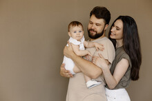 Portrait Of Young Happy Man And Woman Holding Newborn Cute Babe Dressed In White Unisex Clothing. Caucasian Smiling Father And Mother Embracing Tenderly Adorable New Born Child. Happy Family Concept