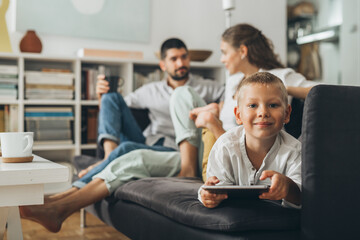 Wall Mural - cute young boy holding tablet computer at home. mother and father in blurred background