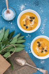 Two white bowls with pumpkin / carrot soup served with cream and pumpkin seeds on blue background, decoreated with sage and salt. Vertical top view.