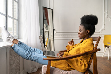 Happy African American woman freelancer with afro hairstyle wear yellow cardigan resting, sitting on chair, legs leaning on windowsill, remote studying, working online on laptop, talking in video chat