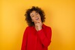 Young arab woman with curly hair wearing red shirt  on yellow background keeps hands partly crossed and hand under chin, looks at camera with pleasure. Happy emotions concept.