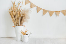 Dry Spikelets Of Pampas Grass In A White Vase And Watering Can Against A White Wall With A Garland Of Burlap Flags.