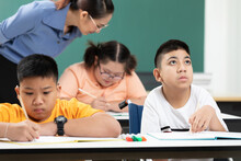 Asian Disabled Kids Or Autism Child Learning Looking And Writing At Desks With Teacher Helping In Classroom