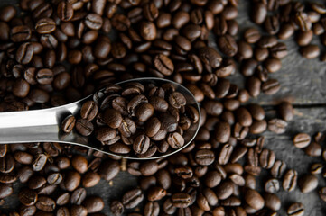 Fresh roasted arabica coffee beans in a metal spoon and scattered coffee beans on a wooden table.