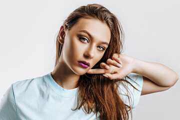 Beautiful young brunette girl with green eyes with long hair with fashion makeup with purple lips on an isolated gray background. The woman holds hand to face and seriously looking at the camera.