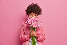Studio Shot Of Good Looking Curly Haired Woman Smells Flowers, Enjoys Pleasant Odor And Stands In Stylish Clothes Indoor. Lovely African American Lady Holds Beautiful Bouquet Of Gerbera Daisy