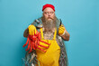 © wayhome.studio  - Thoughtful bearded man holds smoking pipe and octopus, enjoys favorite hobby. Fat successful fisherman with tattooed body, dressed in yellow overalls stands against blue background. Sailor near sea