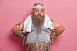 © WHstudio Leushin N - Middle age sad man with thick beard keeps hands on hips and looks aside, takes break after exhausting cardio training. Fatso guy burns calories after nourishing dinner, has sweaty tattooed body