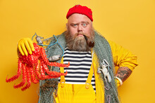 Horizontal Shot Of Self Confident Sailor Has Thick Beard And Holds Big Red Octopus Caught During Fishing. Fisherman Or Angler Poses With Fishing Net, Smokes Tobacco Pipe. Successful Sea Trip