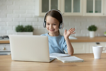 Smiling pretty cute little 7 years old kid girl in headphones looking at laptop screen, waving hello gesture. Happy adorable caucasian child starting distant learning lesson with teacher at home.