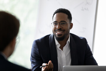 Wall Mural - Smiling African American young businessman in glasses talk with business partner at office meeting. Happy motivated biracial man boss or CEO have talk, discuss cooperation with colleague at briefing.