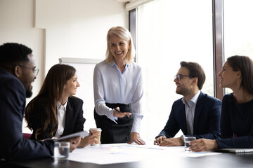 Wall Mural - Smiling middle-aged businesswoman hold lead meeting with diverse colleagues discuss ideas together. Happy multiracial businesspeople have fun brainstorming at team office briefing. Teamwork concept.