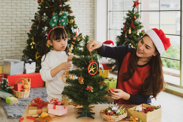 asian mother and her young daughter wearing santa claus hat and using ornament to decorate christmas tree. family together concept