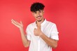 © Roquillo - Young arab man with afro hair wearing shirt standing over isolated red background Showing palm hand and doing ok gesture with thumbs up, smiling happy and cheerful.