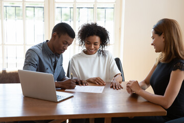 Excited African American husband and wife sit at desk meet with female agent close deal sign agreement, happy biracial couple put signature on contract document consult with realtor in office