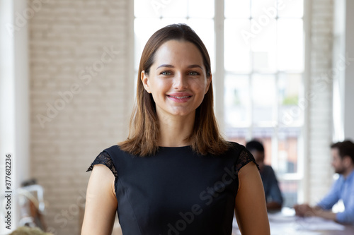 Headshot portrait of smiling millennial businesswoman stand forefront in office look at camera posing, happy confident young Caucasian female employee or company boss CEO show confidence in boardroom