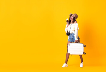 Wall Mural - Cheerful african woman dressed in summer clothes holding passport with ticket while standing with a suitcase over yellow background.