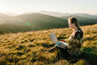© Serhii - Woman traveler using laptop computer while enjoying sunset in mountains. Freelancer girl working on netbook during vacation holidays in autumn. Distant work and travel, freelance as lifestyle concept.