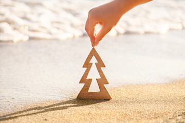 Female hand holding timber wooden Christmas tree on a sand on a tropical beach near ocean, summer Christmas and winter holyday concept, selective focus. Happy New Year background. Zero waste Christmas