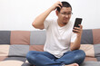 © airdone - Confused Asian man scratch his head when looking at his phone while sitting on sofa, bad news