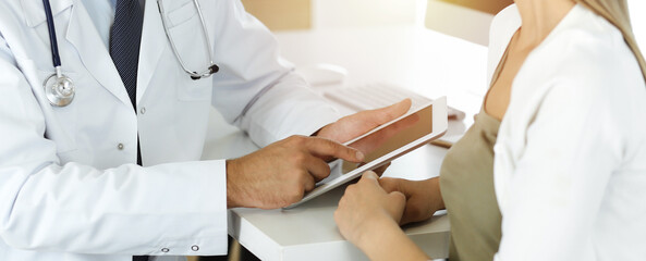 Doctor and patient discussing medical exam results while sitting at the desk in sunny clinic, close-up. Male physician using tablet computer for filling up medication history record.