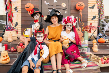 Wall Mural - Young happy woman in witch hat and yellow dress sitting among halloween kids