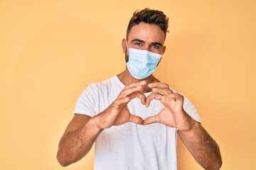 Young hispanic man wearing medical mask smiling in love doing heart symbol shape with hands. romantic concept.