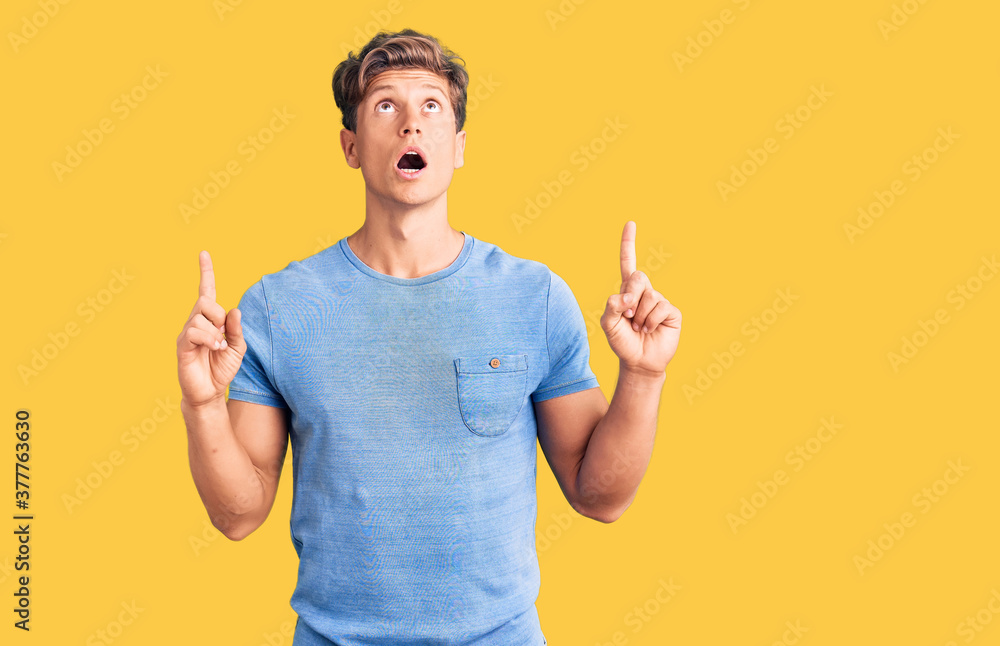 Young handsome man wearing casual clothes amazed and surprised looking up and pointing with fingers and raised arms. - obrazy, fototapety, plakaty 
