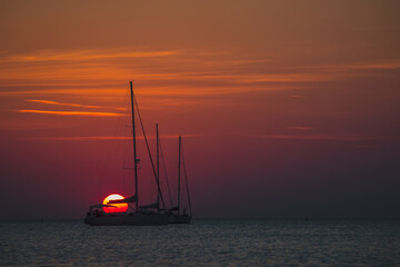 Bloody red sun setting down behind the silohouettes of a two sailboats or yachts moored on an open sea. Adventourous sunset.