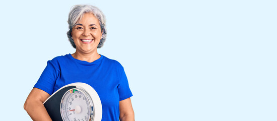 Wall Mural - Senior woman with gray hair holding weight machine to balance weight loss looking positive and happy standing and smiling with a confident smile showing teeth