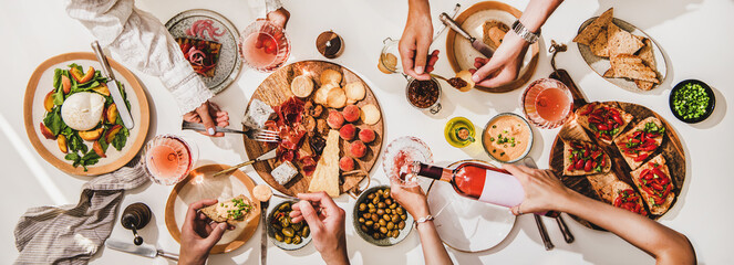 Friends wine and snacks party. Flay-lay of rose wine in glasses, cheese, fruit, meat, tomato brushettas, buratta salad and peoples hands over white table background, top view. Wine tasting concept