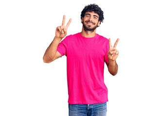 Handsome young man with curly hair and bear wearing casual pink tshirt smiling looking to the camera showing fingers doing victory sign. number two.