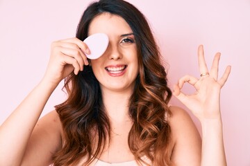 Young beautiful brunette woman holding makeup sponge doing ok sign with fingers, smiling friendly gesturing excellent symbol