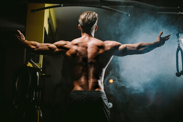 Wall Mural - muscular man posing in gym background. Fitness athlete man showing back muscles in gym. Muscular male shirtless posing with hands up. Black background.