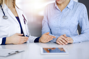 Unknown woman-doctor is showing to her patient a description of medication, while sitting together at the desk in the sunny cabinet in a clinic. Female physician is using a computer tablet and a