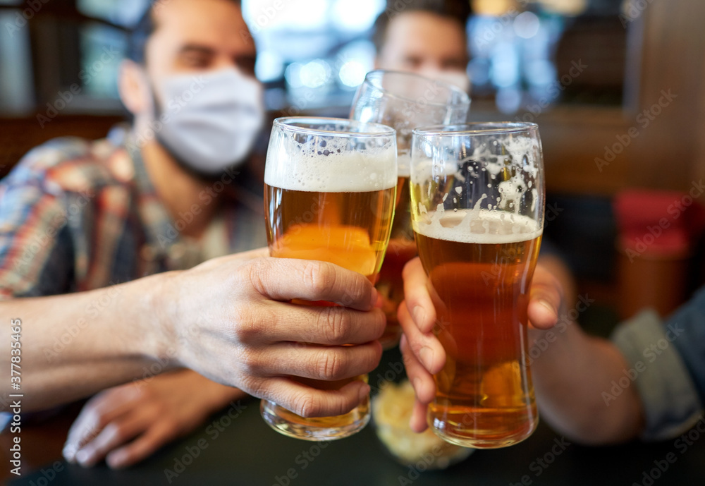 people, leisure and pandemic concept - male friends wearing face protective medical masks for protection from virus disease drinking beer and clinking glasses at bar or pub - obrazy, fototapety, plakaty 