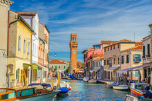 Murano Islands With Clock Tower Torre Dell'Orologio, Boats And Motor Boats In Water Canal, Colorful Traditional Buildings, Venetian Lagoon, Veneto Region, Northern Italy. Murano Postcard Cityscape.