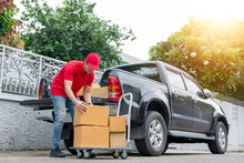 Delivery Men In Red Uniform Unloading Cardboard Boxes From Pickup Truck. Courier Man Sending The Parcel Or Package To The Customer On A Business Day. Online Shopping And Transport Logistics Concept.