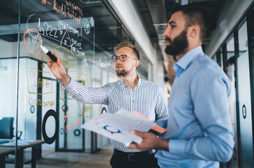 Smart casual men colleagues having productive cooperation on job share ideas during productive conversation in modern workspace with glass board, confident male marketers discussing analytic report