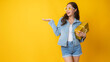 © khwanchai - Portrait of beautiful Asian woman in jean clothes holding gift box and show hand for presentation your product in new year day or birthday, isolated in studio on yellow background
