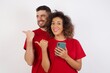 © Roquillo - Portrait of Young beautiful couple wearing red t-shirt on white background using and texting with smartphone  pointing and showing with thumb up to the side with happy face smiling
