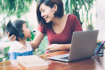 Wall Mural - Young asian mother and son using laptop computer for study and learning together at home, boy wearing headphone for e-learning with distancing, teacher or mom support child, education concept.