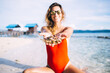 © BullRun - Portrait of funny female swimmer red swimsuit sitting at white sand beach holding collecting mollusk in hands, blurred Caucasian woman enjoying summer vacations for visiting tropical Hawaii