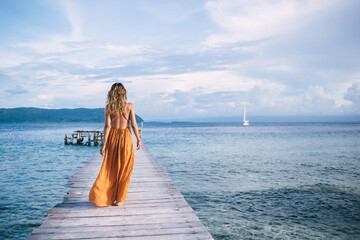 Back view of female tourist enjoying resort vacations in paradise nature environment, woman in stylish sundress walking at pier recreating during solo travelling for visiting Bora Bora island